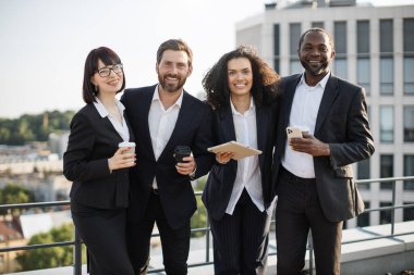 Birlikte poz verip kameraya bakan dost canlısı, çok ahlaklı meslektaşların portresi. Kahveli ve aletli dört neşeli genç insan muhteşem terasta buluşurken gülümsüyor ve eğleniyor..