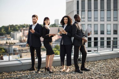 Dört farklı şirket ortağı çatıda duruyor. Şehir manzarası harika ve birbirlerine yaslanıyorlar. İş toplantısı sırasında modern aletler kullanan resmi giyinmiş girişimciler..