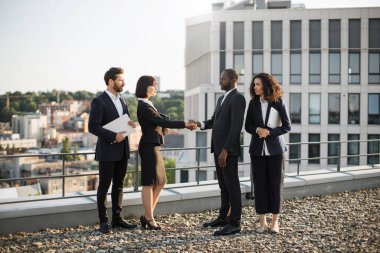 Çift ırklı iş ortakları yeni inşaata karşılıklı yatırım yapılmasını onaylamak için el sıkışıyor. Takım elbiseli dört girişimci ofis terasında toplanıyor. Takım çalışması ve işbirliği kavramı.