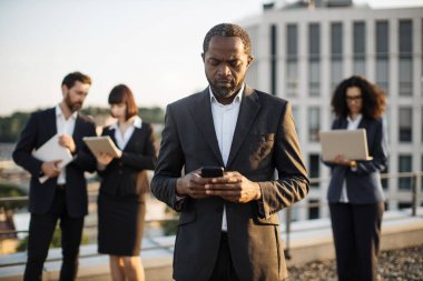 Ciddi yüz ifadesi olan Afrikalı Amerikalı satış müdürü çatıda iş arkadaşlarıyla buluşurken cep telefonundan mesaj okuyor. Emlakçılardan oluşan çeşitli takımlar kişisel cihazlar üzerinde çalışıyor..