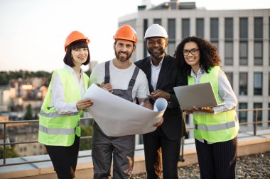 Taşınabilir cihaz ve planlarla kameraya poz veren çok kültürlü geliştiricilerin portresi. Panoramik terasta buluşurken gülümseyen yelekli ve şapkalı dört uluslararası meslektaşım.