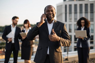 Terasta ortaklarla görüşürken akıllı telefondan Afrikalı Amerikalı beyefendiyi ışınlamak. Takım elbiseli, uzaktan kumandalı aletler kullanan çok ırklı meslektaşlar.