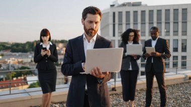 Ofis terasında fantastik şehir manzarasıyla dikilirken dizüstü bilgisayarı olan yakışıklı bir yatırımcı. Uluslararası denetim ekibi yeni proje tasarlamak için aygıtlar kullanıyor.