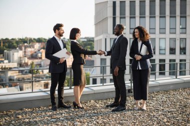 Çift ırklı iş ortakları yeni inşaata karşılıklı yatırım yapılmasını onaylamak için el sıkışıyor. Takım elbiseli dört girişimci ofis terasında toplanıyor. Takım çalışması ve işbirliği kavramı.