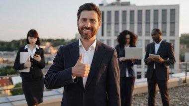 Kameraya gülümseyen, baş parmağını gösteren, iş kıyafeti giymiş özgüvenli sakallı yöneticinin portresi. Melez uyruklu uluslararası meslektaşlar modern cihazların arkasında duruyor..