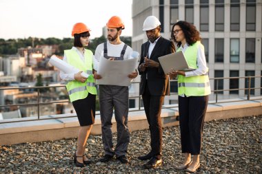 Çok ırklı mimarlar ve tasarımcılar panoramik terasta dikilip proje şemalarına bakıyorlar. Afrika kökenli Amerikalı kadın uzaktan kumandayı tutarken meslektaşları planı işaret ediyor..