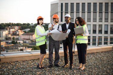 Çok ırklı mimarlar ve tasarımcılar panoramik terasta dikilip proje şemalarına bakıyorlar. Afrika kökenli Amerikalı kadın uzaktan kumandayı tutarken meslektaşları planı işaret ediyor..