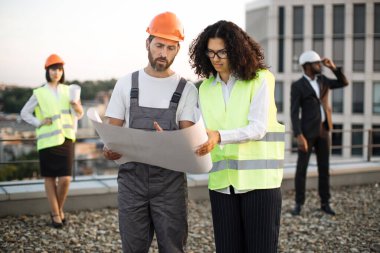 İki mimar inşaat alanını incelerken Afrika kökenli Amerikalı denetçiye teknik planları sunan sert şapkalı bir inşaatçı. Uluslararası müteahhit grubu şehrin çatısında toplandı.