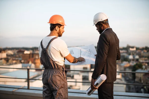 罕见的观点 高加索胡子工人提出的设计蓝图 非洲裔美国城市规划人员在新鲜空气 戴着硬礼帽的男性开发商讨论公寓大楼阳台的位置 — 图库照片