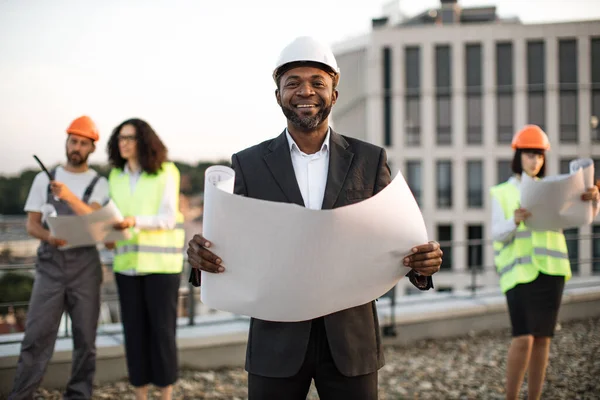 身穿商务服装的非洲裔美国投资者的肖像 在镜头前摆出一副纸制图案 面带微笑的样子 多元文化建筑师小组从天台视察建筑工地 — 图库照片