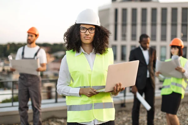 非洲裔美国女性城市规划师的画像 穿着防护服 戴着眼镜 手持远程计算机 看着相机 不同的建筑师评审天台的建筑蓝图 — 图库照片