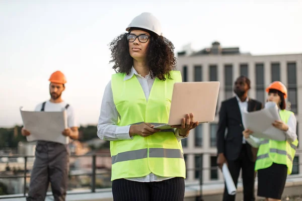 身穿背心 头戴硬礼帽的非裔美国女建筑师手握笔记本电脑站在屋顶上 望着旁边 有蓝图的多种族建筑商在建筑工地观看建筑过程 — 图库照片