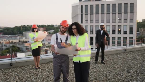 头戴硬礼帽的建筑商在两名建筑师检查建筑工地时 向非洲裔美国主管展示了技术蓝图 国际开发商小组在城市天台开会 — 图库视频影像