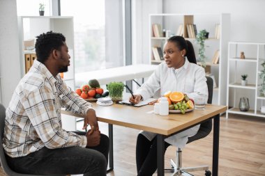 Laboratuvar önlüklü, kendine güvenen kadın diyetisyen klinikteki erkek hastaya danışıyor. Deneyimli bir Afro-Amerikan uzmanı müşterilerine tıbbi geçmişi ve kişisel tercihlerini soruyor..