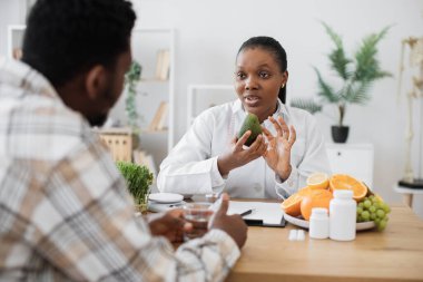 Doktorların işyerinde kalırken laboratuvar önlüklü, yeşil meyveli Afro-Amerikan kadına odaklan. Ciddi bir tıp uzmanı dikkatli bir adamla avokado yemenin faydalarını tartışıyor..