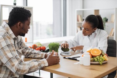 Beyaz önlüklü Afro-Amerikan diyetisyen hasta randevusu sırasında taze avokadoyu beslenme ölçeğine koyuyor. Kadın profesyonel akıllı teknolojilerle kalorilerin doğru ölçümlerini alıyor..