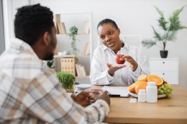 Taze elmayla sağlık uzmanına yakın durun ve danışma odasında bireyle konuşurken ölçü alın. Laboratuvar önlüğü danışmanlığı yapan çok ırklı bir kadın genel sağlık ve kapalı alanlarda refah konusunda.