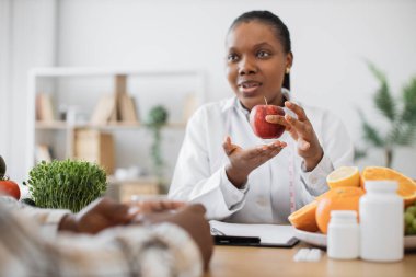Taze elmayla sağlık uzmanına yakın durun ve danışma odasında bireyle konuşurken ölçü alın. Laboratuvar önlüğü danışmanlığı yapan çok ırklı bir kadın genel sağlık ve kapalı alanlarda refah konusunda.