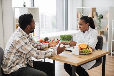 Kendine güveni tam bir tıp uzmanı. Ofis masasında oturan sıradan giysili yetişkin bir adama mikro yeşillikli konteynır gösteriyor. Çok kültürlü beslenme uzmanları sağlıklı beslenmeye iyi katkı öneriyor..