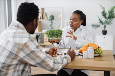 Kendine güveni tam bir tıp uzmanı. Ofis masasında oturan sıradan giysili yetişkin bir adama mikro yeşillikli konteynır gösteriyor. Çok kültürlü beslenme uzmanları sağlıklı beslenmeye iyi katkı öneriyor..