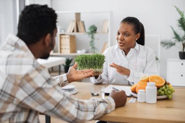 Kendine güveni tam bir tıp uzmanı. Ofis masasında oturan sıradan giysili yetişkin bir adama mikro yeşillikli konteynır gösteriyor. Çok kültürlü beslenme uzmanları sağlıklı beslenmeye iyi katkı öneriyor..
