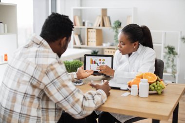 Doktorların iş yerindeki çok kültürlü bir adam için tabletle beslenme verilerini grafiksel olarak gösteren deneyimli bir profesyonel. Diyet uzmanı kadın müşteriyi tedavi sırasında doğru yiyecek seçimlerine motive ediyor.