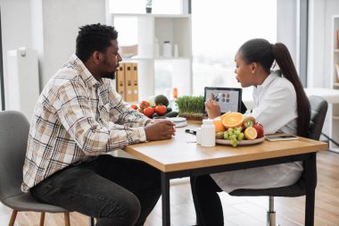 Afro-Amerikan kadının, hastaların beslenme verilerini muayene odasında dijital tabletle analiz edişinin omuzlarının üstünden görünüşü. Tıp merkezindeki erkek hastaların sağlık ihtiyaçlarını değerlendirmek için etkili bir dietisyen..