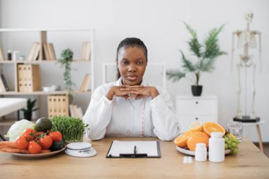 Klinikte profesyonel faaliyet gösteren doktor önlüklü çok ırklı ciddi bir kadın beslenme uzmanı. Ofisteki müşterilerin yemek planları üzerine kendinden emin sağlık uzmanı düşüncesi.