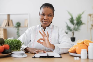 Doktor önlüklü çekici yetişkin kadın portresi üzerinde sağlıklı yiyecek ve ilaç ile ofis masasında poz veriyor. Çok kültürlü diyet planlaması ve işaretleri gösteren beslenme programları.