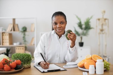 Elinde avokado olan beyaz önlüklü, yazı masasında poz veren gülümseyen çok kültürlü kadının portresi. Neşeli bir yiyecek ve beslenme uzmanı. Doktorların iş yerinde özel diyet için yiyecek seçiyor..