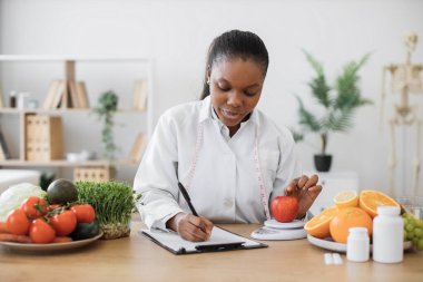 Laboratuvar önlüklü çok ırklı bir kadın elmayı diyet ölçeğinde muayene odasında değerlendirirken not alıyor. Beslenmeyi keyifli tutmak için ikame hizmetleri sunan düşünceli bir sağlık uzmanı.