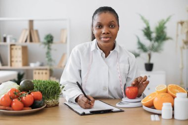 Laboratuvar önlüklü çok ırklı bir kadın elmayı diyet ölçeğinde muayene odasında değerlendirirken not alıyor. Beslenmeyi keyifli tutmak için ikame hizmetleri sunan düşünceli bir sağlık uzmanı.