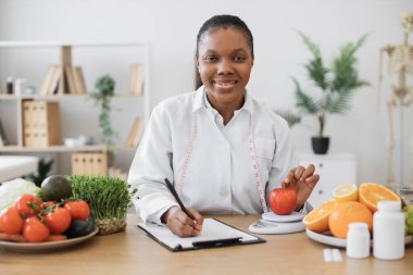 Laboratuvar önlüklü çok ırklı bir kadın elmayı diyet ölçeğinde muayene odasında değerlendirirken not alıyor. Beslenmeyi keyifli tutmak için ikame hizmetleri sunan düşünceli bir sağlık uzmanı.