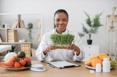 Çalışma masasında otururken beyaz önlüklü, elinde mikro yeşillik kabıyla çekici bir bayanın yakın görüntüsü. Afrika kökenli Amerikalı beslenme uzmanı diyete modern ekleme öneriyor.