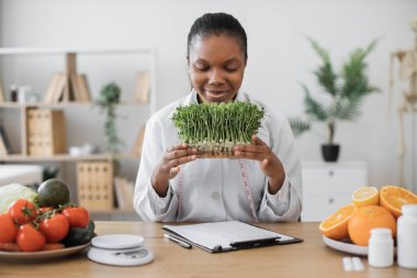 Özel kliniğin danışma odasında gülümseyen mikroyeşilliklere bakan çok ırklı doktor önlüklü çekici bir kadın. Deneyimli dietisyen yiyeceğe farklı vitaminler, mineraller ve besinler ekliyor..