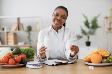 Ofisteki çok kültürlü bir kadın tarafından tutulan vitamin takviyeli ilaç kutularına odaklan. Beyaz önlüklü tıp uzmanı beslenme boşluklarını doldurmakla ilgili tavsiyelerde bulunuyor..