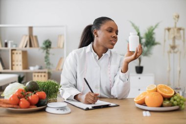 Ciddi bir Afro-Amerikalı kadın klinikteki yazı masasında not alırken ilaç kutusunda etiket okuyor. Doktor önlüğü konusunda diyet uzmanı şişe içeriğinin besleyici gerçeklerini inceliyor.