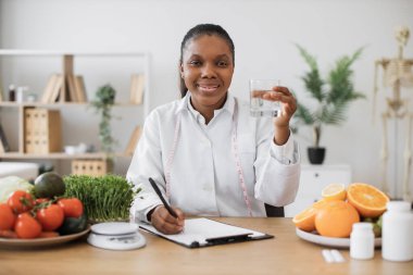Beyaz önlüklü, odaklanmış Afro-Amerikan kadın doktorların iş yerindeki camlarda sıvı temizliğini kontrol ediyor. Etkili beslenme uzmanı diyette metabolizmayı geliştirmek için içme suyu hakkında tavsiye veriyor..
