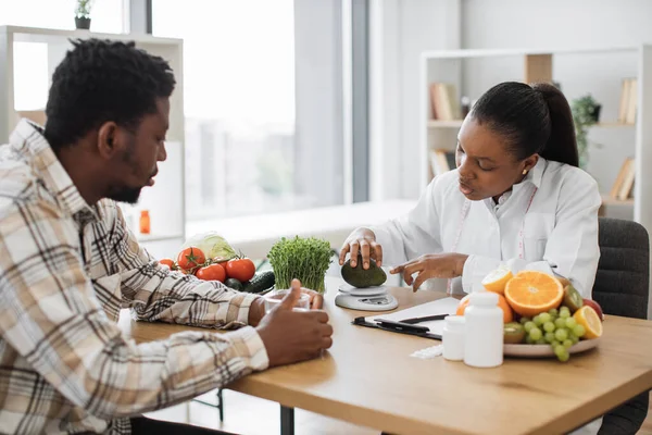 Beyaz önlüklü Afro-Amerikan diyetisyen hasta randevusu sırasında taze avokadoyu beslenme ölçeğine koyuyor. Kadın profesyonel akıllı teknolojilerle kalorilerin doğru ölçümlerini alıyor..