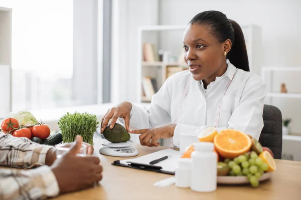 白いコートを着たアフリカ系アメリカ人の栄養士は 患者の任命中に栄養スケールに新鮮なアボカドを置きます スマート技術でカロリーの正確な測定を取得する女性の専門家 — ストック写真