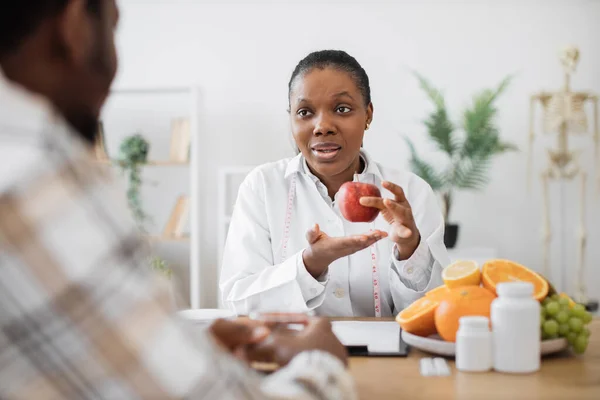 プロのオフィスで男性の大人にリンゴを示すテープを測定するアフリカ系アメリカ人の栄養士に焦点を当てます 医師の信頼できる専門家は スポーツ栄養食品を消費する動機をコート — ストック写真