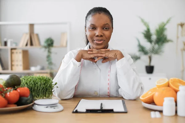 医師の深刻な多民族の女性は 診療所で栄養専門家としての専門活動を行うコート オフィスの机で顧客の食事計画についての健康の専門家の思考を確信しなさい — ストック写真