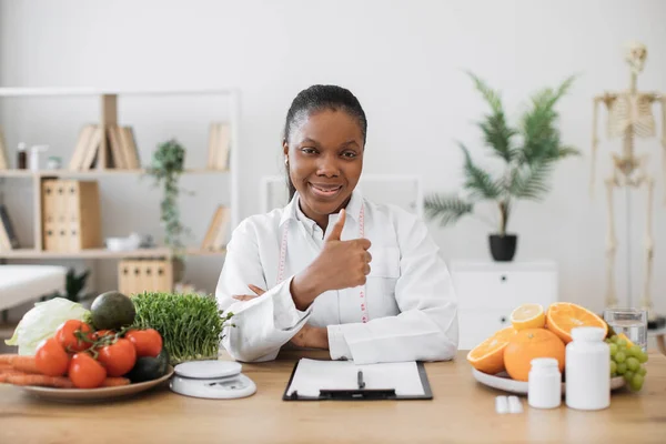 医師の魅力的な大人の女性の肖像は それに健康的な食べ物や薬でオフィスの机の上にポーズコート 多文化食生活計画と親指を示す栄養プログラムを実施 — ストック写真