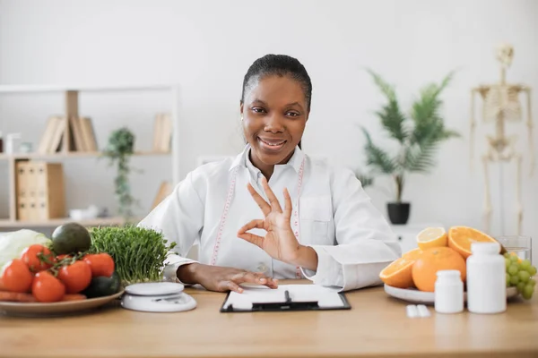 医師の魅力的な大人の女性の肖像は それに健康的な食べ物や薬でオフィスの机の上にポーズコート 多文化食の企画 運営栄養プログラムサインOk — ストック写真