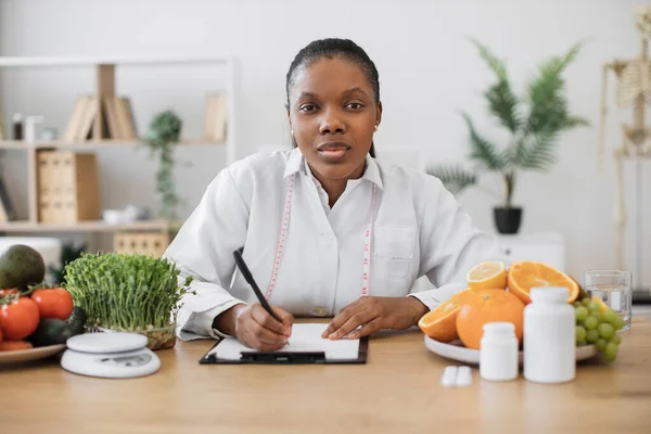 オフィスの筆記机に座っている間 クリップボードの上にペンを保持している研究室のコートにアフリカ系アメリカ人女性の肖像画 居心地の良い職場での患者の食事計画の変化を作る栄養専門家 — ストック写真
