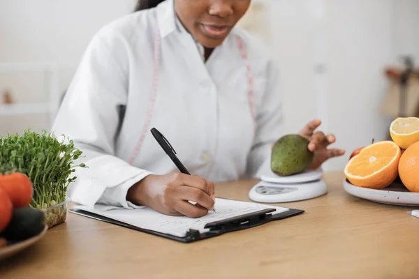 栄養スケールで緑の果実を持つ多人種女性の作物ビューでは オフィスに詳細を置きながら 仕事中の食品に関する栄養情報を特定する経験豊富な医療専門家 — ストック写真