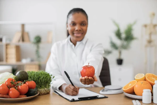 白いコートを着たアフリカ系アメリカ人女性が笑顔で手に持っている熟した赤いリンゴに焦点を当てる屋内 健康的な食事習慣を教えることにより 健康状態を管理する栄養学の専門家 — ストック写真