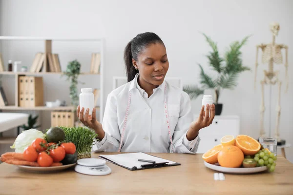 女性栄養士に焦点を当て 2本の薬瓶を持ちながら オフィスのデスクで製品を扱っています 顧客の食事のバランスをとるための食品サプリメントを選択する多民族健康専門家 — ストック写真
