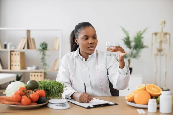 白いコートを着たアフリカ系アメリカ人女性に焦点を当て 医師の職場でガラスの流動的な透明度をチェックします 食事における代謝を促進するための飲料水に関する効果的な栄養専門家の助言 — ストック写真
