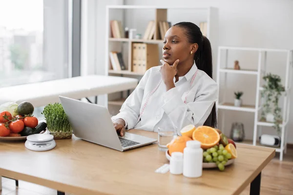 Serious multicultural lady in doctors coat typing on modern laptop in consulting room of medical center. Experienced nutrition professional searching for weight loss information on food websites.